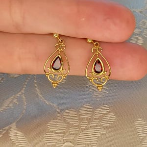 K&ouml;nnte beinhalten: Ein Paar goldfarbene Ohrh&auml;nger. Jeder Ohrring hat einen tropfenf&ouml;rmigen, dunkelroten Edelstein, der in einem verzierten Filigranmuster gefasst ist. Die Ohrringe haben eine zarte, Vintage-&Auml;sthetik.