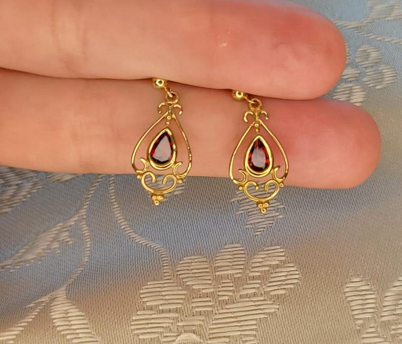 K&ouml;nnte beinhalten: Ein Paar goldfarbene Ohrh&auml;nger. Jeder Ohrring hat einen tropfenf&ouml;rmigen, dunkelroten Edelstein, der in einem verzierten Filigranmuster gefasst ist. Die Ohrringe haben eine zarte, Vintage-&Auml;sthetik.