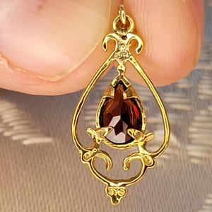K&ouml;nnte beinhalten: Goldfarbener Anh&auml;nger mit einem tropfenf&ouml;rmigen, dunkelroten Edelstein. Der Anh&auml;nger hat ein verziertes, offenes Design mit dekorativen Verzierungen. Der Edelstein ist facettiert und f&auml;ngt das Licht ein. Der Anh&auml;nger ist an einer kleinen Schlaufe befestigt.