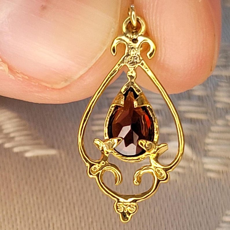 K&ouml;nnte beinhalten: Goldfarbener Anh&auml;nger mit einem tropfenf&ouml;rmigen, dunkelroten Edelstein. Der Anh&auml;nger hat ein verziertes, offenes Design mit dekorativen Verzierungen. Der Edelstein ist facettiert und f&auml;ngt das Licht ein. Der Anh&auml;nger ist an einer kleinen Schlaufe befestigt.