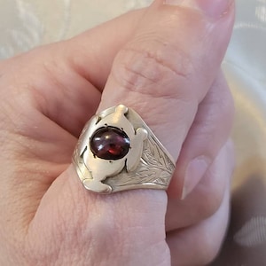 Könnte beinhalten: Ein silberner Ring mit einem roten Edelstein, der in einem stilisierten Design gefasst ist. Der Ring ist mit einem floralen Muster graviert.