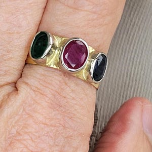 Puede incluir: Un anillo de oro con tres piedras preciosas de forma ovalada. La piedra central es un rubí rojo oscuro, flanqueado por una esmeralda verde oscuro a la izquierda y un zafiro azul oscuro a la derecha.