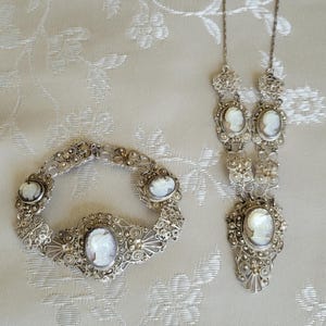 Könnte beinhalten: Ein silberfarbenes Schmuckset mit Cameo-Anhängern. Das Set umfasst eine Halskette und ein Armband, beide mit ovalen Cameo-Designs. Armband und Halskette sind auf einem cremefarbenen Stoff mit Blumenmuster ausgestellt.
