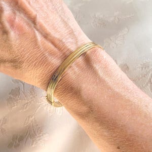 May include: A gold-colored bangle bracelet with three parallel bands. The bracelet is secured with a clasp. The jewelry is worn on a wrist, and the background is a light-colored fabric with a floral pattern.