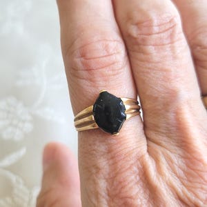 Könnte beinhalten: Ein goldfarbener Ring mit einem dunklen, unregelmäßig geformten Stein. Der Ring hat ein dreifaches Banddesign, wobei der Stein prominent in der Mitte platziert ist. Der Ring ist ein Schmuckstück.