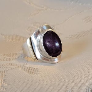 May include: A silver ring featuring a large, oval, dark purple gemstone. The ring has a modern design with a wide band and a bezel setting that securely holds the stone. The ring is displayed on a textured surface.