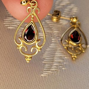 K&ouml;nnte beinhalten: Ein Paar goldfarbene Ohrringe mit einem tropfenf&ouml;rmigen, tiefroten Edelstein in der Mitte. Die Ohrringe haben ein verziertes Filigran-Design mit dekorativen Wirbeln und einem Steckerverschluss. Die Ohrringe werden vor einem neutralen Hintergrund pr&auml;sentiert.
