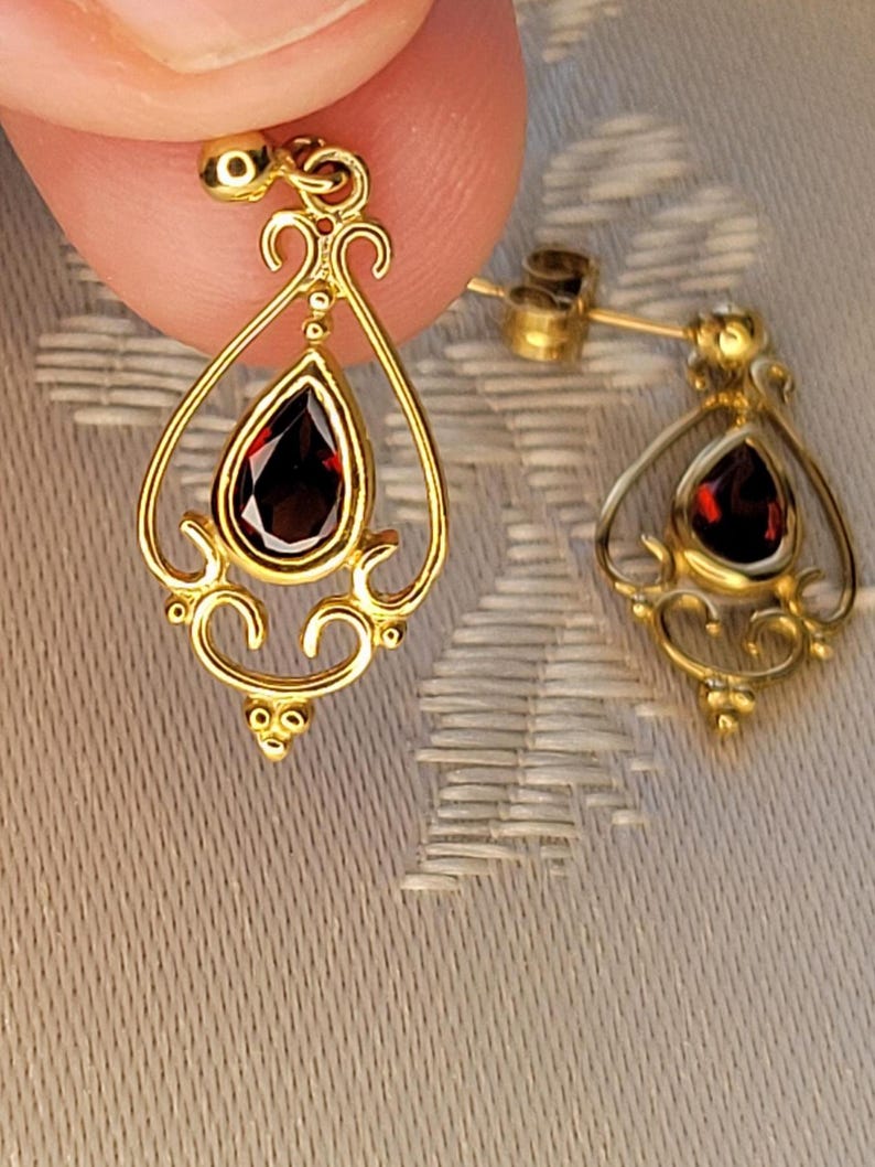 K&ouml;nnte beinhalten: Ein Paar goldfarbene Ohrringe mit einem tropfenf&ouml;rmigen, tiefroten Edelstein in der Mitte. Die Ohrringe haben ein verziertes Filigran-Design mit dekorativen Wirbeln und einem Steckerverschluss. Die Ohrringe werden vor einem neutralen Hintergrund pr&auml;sentiert.