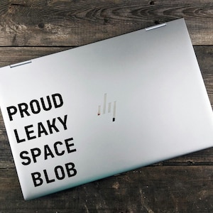 May include: A silver laptop with a metallic finish featuring a black decal on the left side that reads PROUD LEAKY SPACE BLOB in a bold sans-serif font. The laptop lid displays a centered metallic logo consisting of four vertical lines.