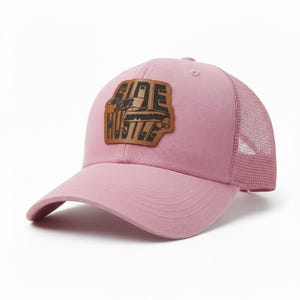 Op de afbeelding: Roze baseballpet met een mesh achterkant en een gebogen klep. De pet heeft een bruin leren patch met de woorden "SIDE HUSTLE" in een vet, gestileerd lettertype. De pet is geschikt voor casual wear.
