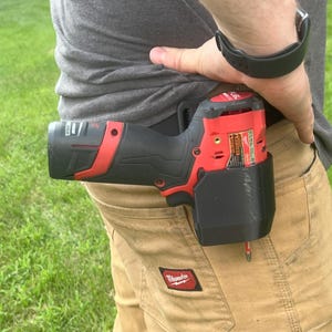 May include: A red and black cordless drill is secured in a black holster attached to a tan work shorts pocket. The drill is a power tool, likely for construction or DIY projects. The shorts have a small red logo.