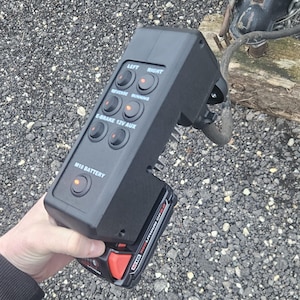 May include: A black control panel with multiple illuminated buttons, labeled "LEFT," "RIGHT," "REVERSE," "RUNNING," "E-BRAKE 12V AUX," and "M18 BATTERY." The panel is connected to a trailer hitch and powered by a red and black battery.