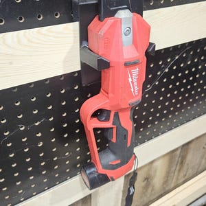 May include: A red and black Milwaukee M12 cordless cable cutter mounted on a black pegboard. The tool is held in place by a black metal bracket. The pegboard has evenly spaced holes. A black strap hangs from the tool.