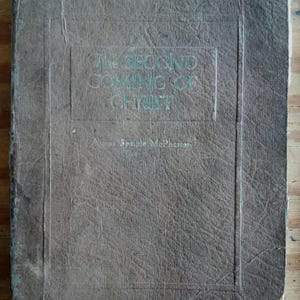 Può includere: Un libro marrone usurato con il titolo "The Second Coming of Christ" in lettere verdi in rilievo. La copertina è strutturata e mostra segni di età, con il nome dell'autore, Aimee Semple McPherson, in basso.