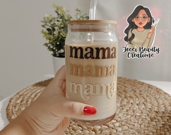 May include: A clear glass tumbler with a brown wooden lid and a straw. The tumbler has the word "mama" written on it three times in different colors and fonts. The tumbler is being held by a hand.