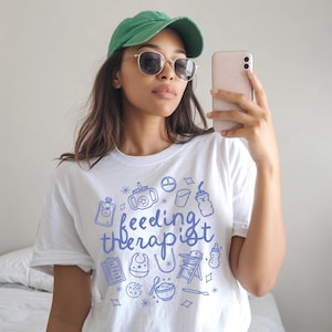 May include: White t-shirt with the words "feeding therapist" in blue script and baby-related illustrations. The person in the image is wearing a green cap and sunglasses.