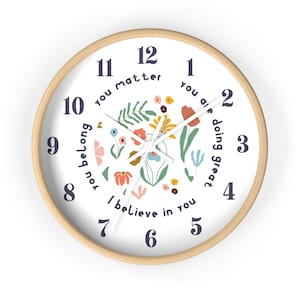 May include: A white round clock with a wooden frame and floral design. The clock face has black numbers and the words "you matter", "you belong", "you are doing great", and "I believe in you" written around the numbers.