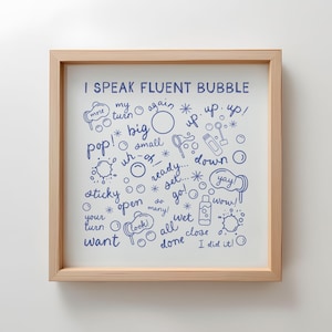 May include: A framed print with the text "I SPEAK FLUENT BUBBLE" in blue, surrounded by bubble-themed doodles and words like "pop," "yay," and "wow." The frame is made of light-colored wood.