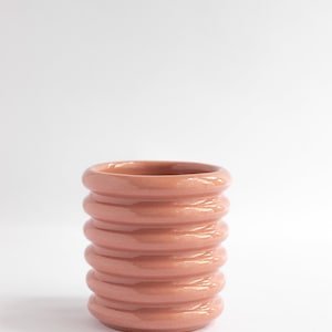 Puede incluir: Un jarrón de cerámica color terracota con un diseño acanalado. El jarrón tiene forma cilíndrica con anillos redondeados y apilados. El acabado brillante refleja la luz, resaltando la textura. Adecuado para la decoración del hogar.