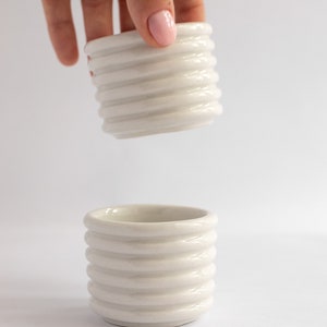 Puede incluir: Dos tazas de cerámica blanca con un diseño acanalado. Las tazas están apiladas una encima de la otra.