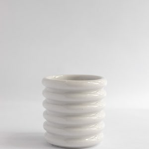 Puede incluir: Un pequeño jarrón de cerámica blanca con un diseño acanalado. El jarrón tiene forma cilíndrica con crestas redondeadas que rodean el cuerpo. El diseño minimalista y el color neutro lo hacen adecuado para varios estilos de decoración.