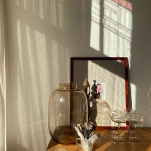 Op de afbeelding: Een stilleven met een grote, bruine glazen vaas, een kleine decoratieve beker met veren en verschillende wijnglazen. Een ingelijste foto en andere decoratieve flessen zijn ook aanwezig op een houten oppervlak, verlicht door zonlicht.