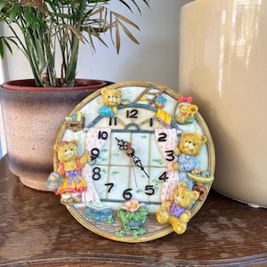 Puede incluir: Un reloj redondo decorativo con un diseño caprichoso de osito de peluche. La esfera del reloj tiene un fondo claro con números y manecillas negras. El reloj está rodeado de figuras de ositos de peluche con varios atuendos. El reloj está sobre una superficie de madera.
