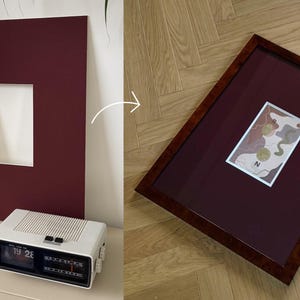 May include: A framed artwork with a burgundy mat and a dark wood frame. The artwork features abstract shapes in neutral tones with gold accents. A vintage white alarm clock is in the foreground.