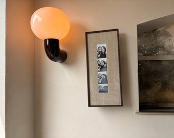 Può includere: Un'applique da parete con una lampadina rotonda arancione brillante e un braccio nero curvo. Una striscia di foto incorniciata con immagini in bianco e nero è appesa al muro. Il muro è dipinto di beige chiaro.