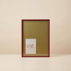 Op de afbeelding: Een rechthoekige fotolijst met een dieprode rand en een goudkleurige passe-partout. Binnenin de lijst bevindt zich een klein wit rechthoekig inzetstuk met de letter R in een decoratief, donkerrood cursief handschrift in het midden.