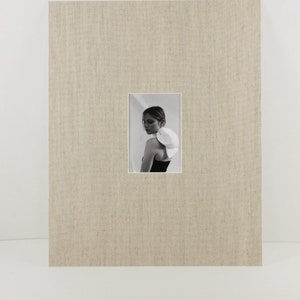 Pode incluir: Fotografia a preto e branco de uma pessoa com um top preto e um acessório escultural branco. A fotografia está dentro de uma moldura retangular, montada sobre um fundo de tecido bege texturizado.