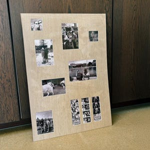 Op de afbeelding: Een houten fotodisplaybord met verschillende zwart-witfoto's van verschillende formaten. De foto's zijn gerangschikt op een lichtgekleurd houten oppervlak en leunen tegen een donkerbruine kast.