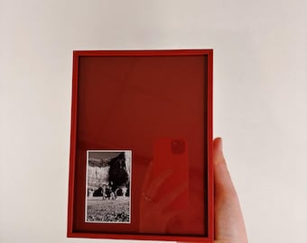 Könnte beinhalten: Ein roter, rechteckiger Bilderrahmen mit einem Schwarz-Weiß-Foto. Der Rahmen wird von einer Hand gehalten. Das Foto zeigt Menschen vor einer Felsformation. Der Hintergrund ist weiß.