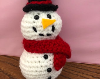 May include: A crocheted snowman with a black top hat, red scarf, and black button eyes. The snowman has an orange carrot nose.