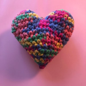 Könnte beinhalten: Ein handgefertigtes, mehrfarbiges Häkelherz. Das Herz ist aus Garn in den Farbtönen Rosa, Blau, Grün und Gelb gefertigt. Das Herz befindet sich auf einem rosa Hintergrund. Oben ist eine kleine silberne Metallschlaufe angebracht.