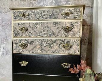 May include: A black chest of drawers with gold hardware and a floral patterned design on the top three drawers. The chest of drawers is sitting on a wooden floor.