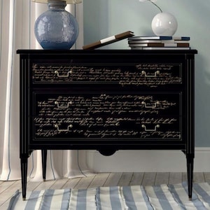 May include: Black three-drawer chest with gold-toned handles and script detailing. The chest has a classic design with slender legs and a curved apron. A blue glass lamp and books are on top.