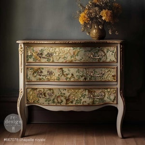 Puede incluir: C&oacute;moda de tres cajones con dise&ntilde;o floral. La c&oacute;moda est&aacute; pintada en color gris claro con detalles dorados y herrajes ornamentados. Los cajones presentan un detallado estampado floral en tonos verdes, rosas y dorados. Un jarr&oacute;n con flores amarillas encima.