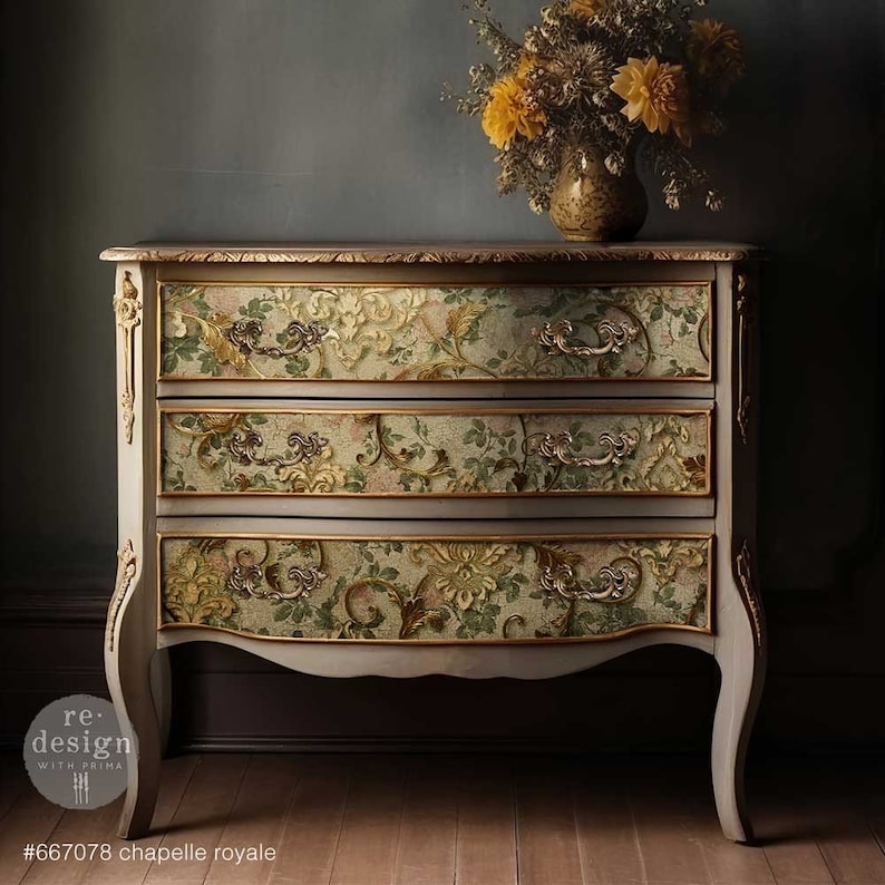 Puede incluir: C&oacute;moda de tres cajones con dise&ntilde;o floral. La c&oacute;moda est&aacute; pintada en color gris claro con detalles dorados y herrajes ornamentados. Los cajones presentan un detallado estampado floral en tonos verdes, rosas y dorados. Un jarr&oacute;n con flores amarillas encima.