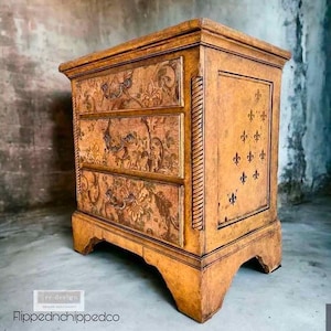 Puede incluir: Una mesita de noche de madera de tres cajones con acabado envejecido. Los cajones presentan dise&ntilde;os florales y tiradores ornamentados. El panel lateral muestra dise&ntilde;os de flor de lis. La marca es "Flippednchippedco".