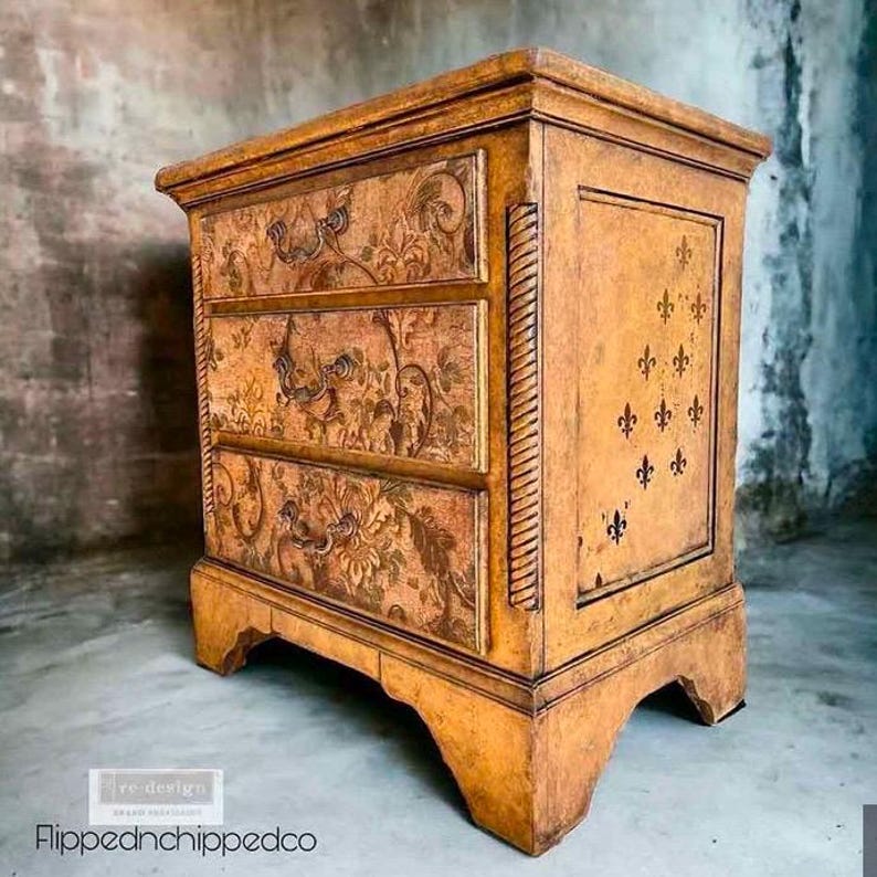 Puede incluir: Una mesita de noche de madera de tres cajones con acabado envejecido. Los cajones presentan dise&ntilde;os florales y tiradores ornamentados. El panel lateral muestra dise&ntilde;os de flor de lis. La marca es "Flippednchippedco".