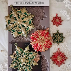 May include: A package of Decor Moulds with the text "FROSTED SNOWFLAKE WONDERS". The package includes a mold and several finished snowflake designs in gold, green, and red. The mold measures 12.7cm x 25.4cm.