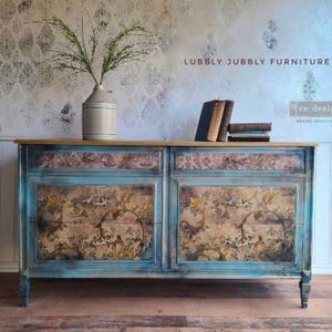 Puede incluir: Una c&oacute;moda azul y marr&oacute;n desgastada con frentes de cajones con estampado floral y una tapa de madera clara. La c&oacute;moda tiene herrajes ornamentados y est&aacute; decorada con un jarr&oacute;n con vegetaci&oacute;n y libros. El texto "LUBBLY JUBBLY FURNITURE" es visible.