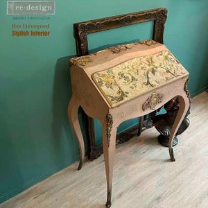 Puede incluir: Un escritorio de escritura de estilo vintage con una parte superior inclinada y patas curvas. El escritorio est&aacute; pintado en un tono rosa suave, con adornos dorados ornamentados y un dise&ntilde;o floral en la parte superior. El texto "re-design" y "Ilse Lievegoed Stylish Interior" es visible.