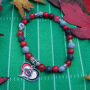 Könnte beinhalten: Ein Armband mit roten, grauen und dunkelblauen Perlen, akzentuiert mit silberfarbenen Abstandshaltern. Ein herzförmiger Anhänger mit dem Ohio State-Logo hängt daran. Das Armband wird auf einer grünen Oberfläche präsentiert.