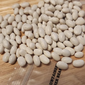 May include: A pile of dried, white beans, possibly cannellini or navy beans, are scattered on a light brown wooden surface. The beans are oval-shaped and have a smooth texture. The image is well-lit, highlighting the natural color and shape of the beans.