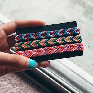 Könnte beinhalten: Drei bunte Freundschaftsbänder mit geometrischen Mustern auf schwarzem Hintergrund. Die Armbänder sind aus buntem Stickgarn gefertigt.