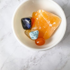 May include: A white bowl filled with various crystals, including a large orange calcite, a blue sodalite, a yellow citrine, a blue and green angelite, and a small orange carnelian.