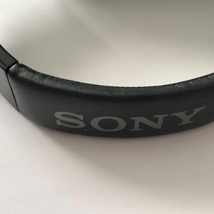 May include: Close-up of a black headphone headband with the brand name "SONY" printed in gray. The headband is made of a smooth, dark material and is curved. The background is white.