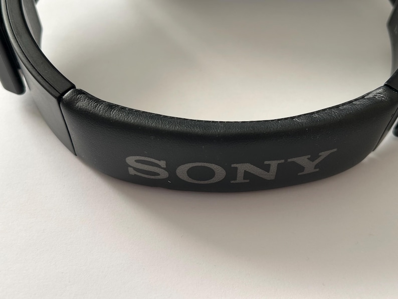May include: Close-up of a black headphone headband with the brand name "SONY" printed in gray. The headband is made of a smooth, dark material and is curved. The background is white.